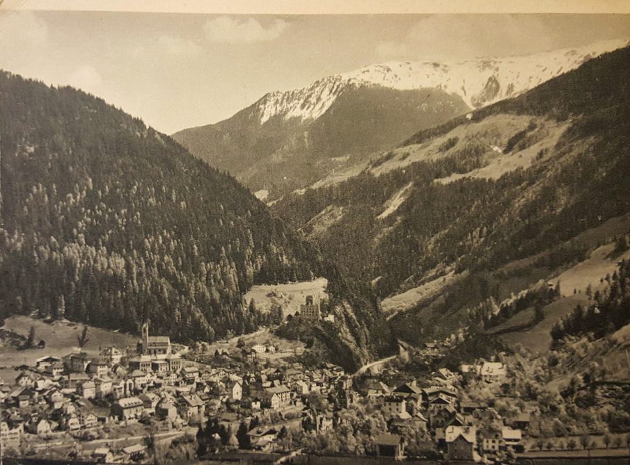 A sepia photograph postcard of Landeck in the Tyrol: the town fills the valley floor, a church spire rises at the left, a castle stands on a wooded spur in the center, and snow-covered mountains rise behind.