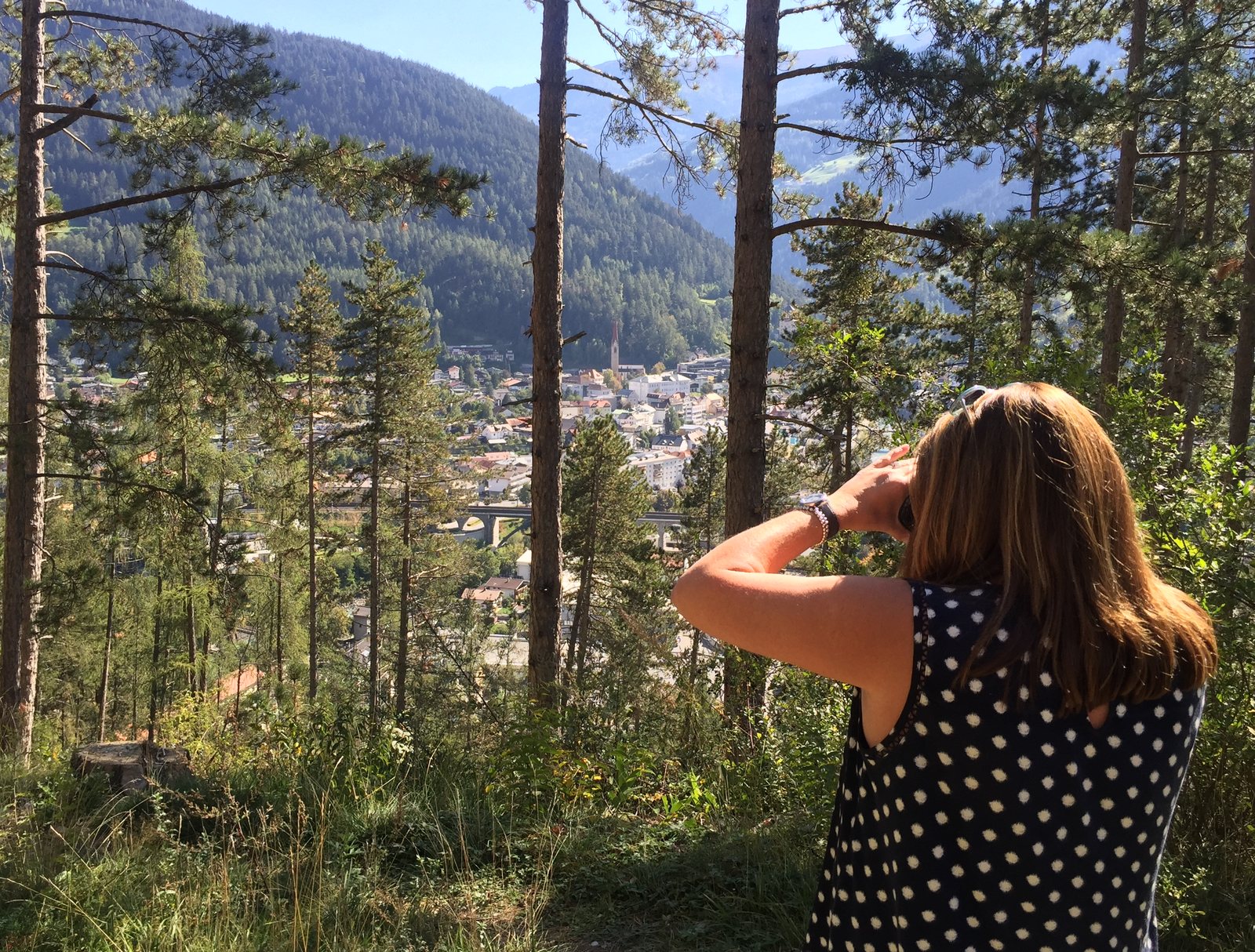 A woman with long brown hair, seen from behind among pine trees on a hillside, raises a camera toward the valley below.