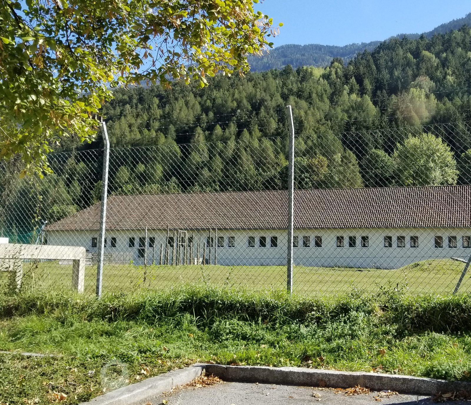 Long, low barracks buildings with white walls and a brown tiled roof behind a chain-link fence, wooded mountainside rising behind, a tree in the foreground.