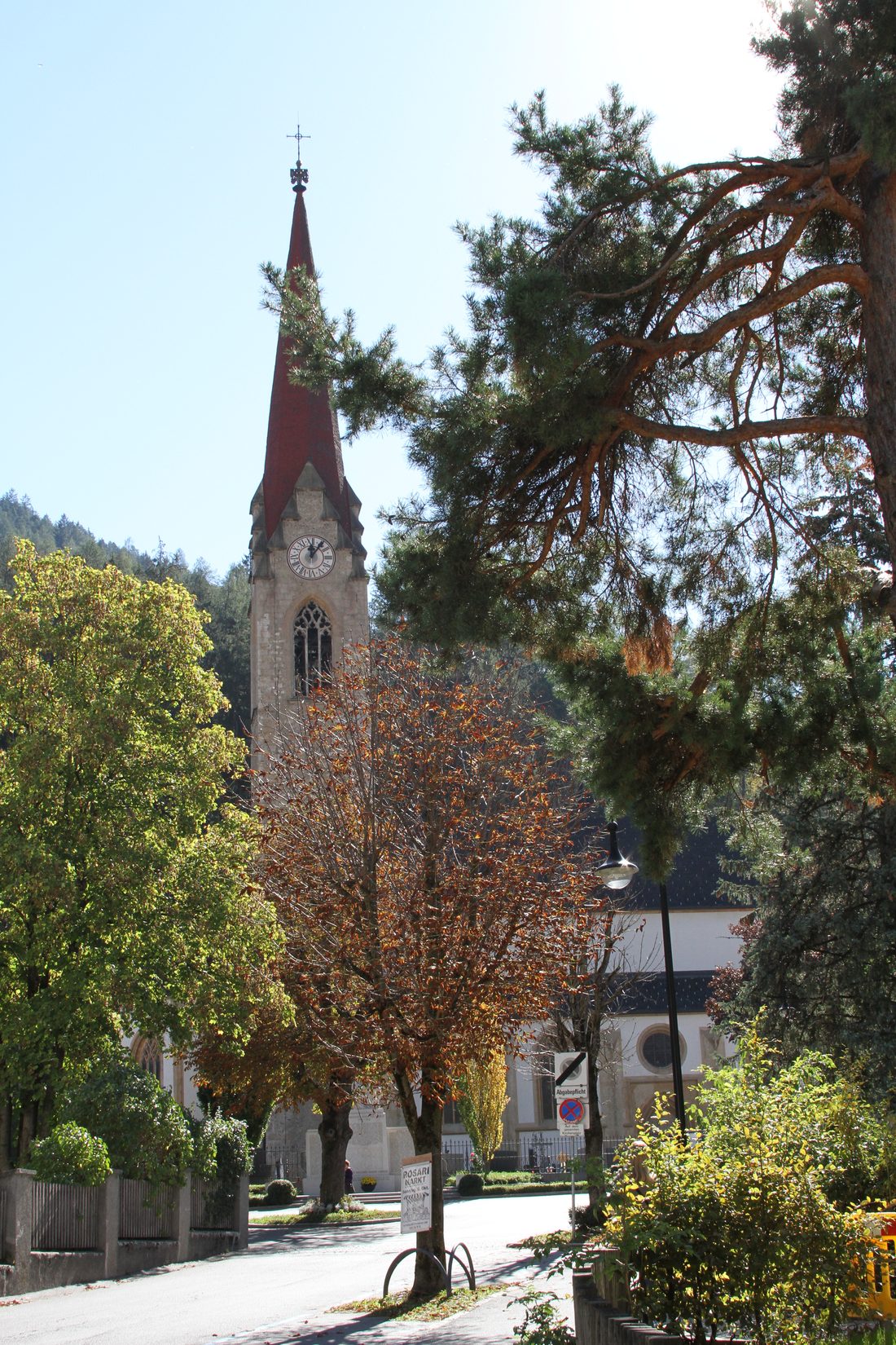 The parish church of Landeck seen through autumn trees, a tall stone tower with a clock face and a steep red spire against a bright sky.