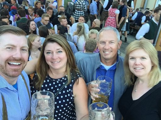 Four friends crowded together in a packed Oktoberfest beer hall, smiling at the camera and holding up liter glasses of beer.
