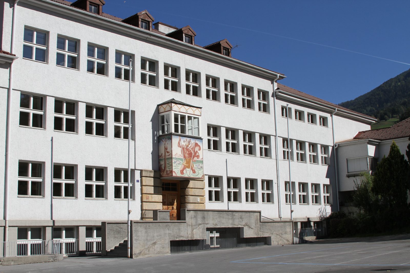A long three-story school building in Landeck, white with rows of square windows, a painted oriel bay over the entrance and a paved yard in front, under a clear blue sky.