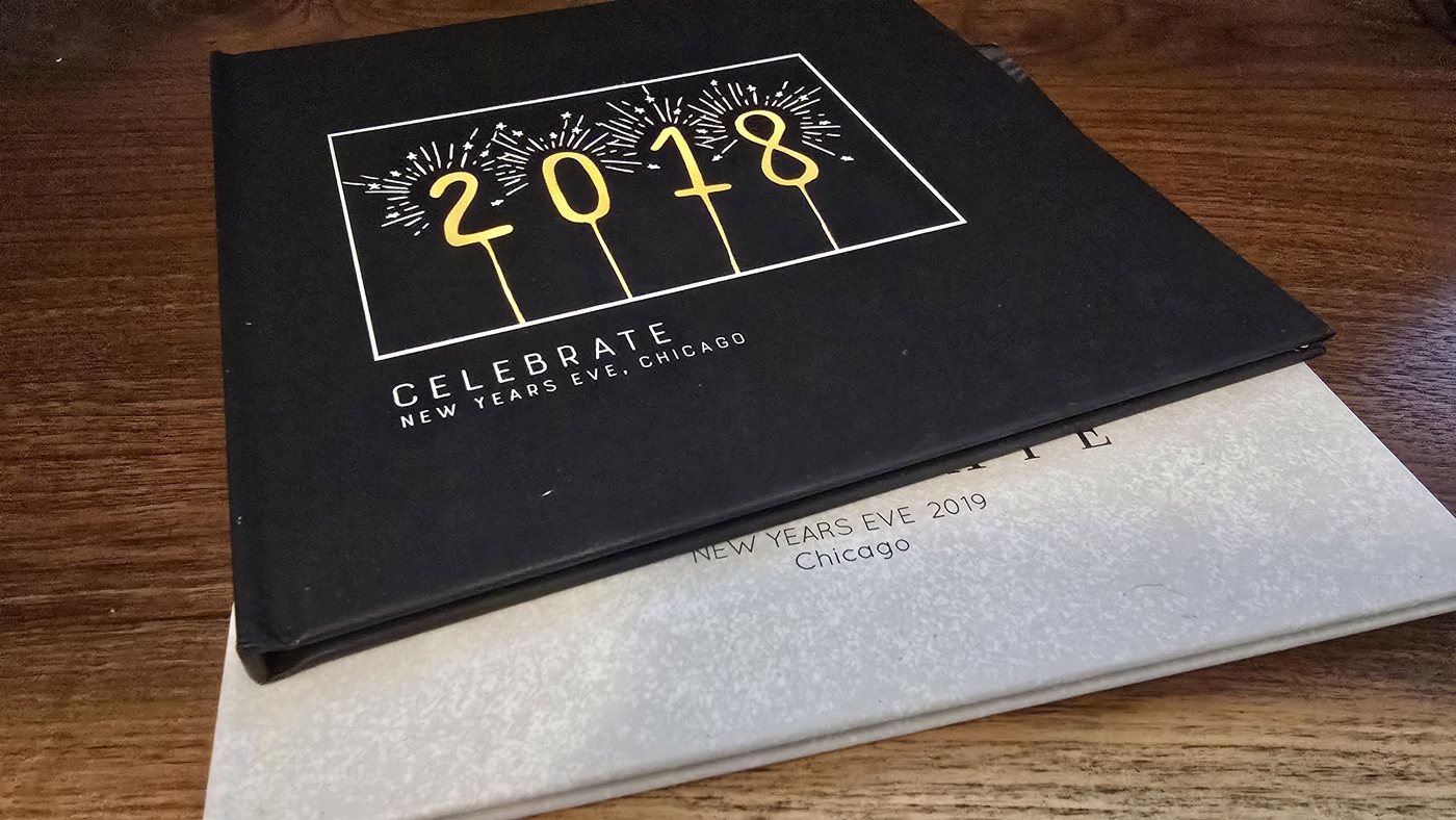 Two Milestone books stacked on a wood table. The top volume is black cloth, blocked in white and gold with sparkler numerals reading 2018 and the words Celebrate, New Years Eve, Chicago. Beneath it a pale gray volume is blind-blocked New Years Eve 2019, Chicago.