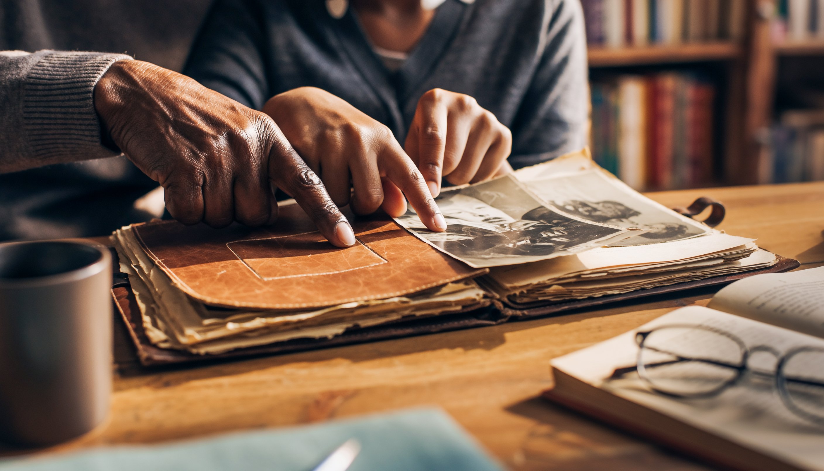 Two people at a wooden table point together at old black-and-white photographs in a worn leather album, one hand older and one younger. A mug, a pair of reading glasses resting on an open book, and shelves of books sit around them.